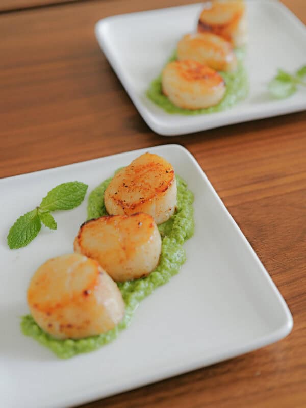 Three seared scallops are served on a bed of green puree, garnished with a mint sprig, on a white square plate. Another similar plate is blurred in the background on a wooden table.
