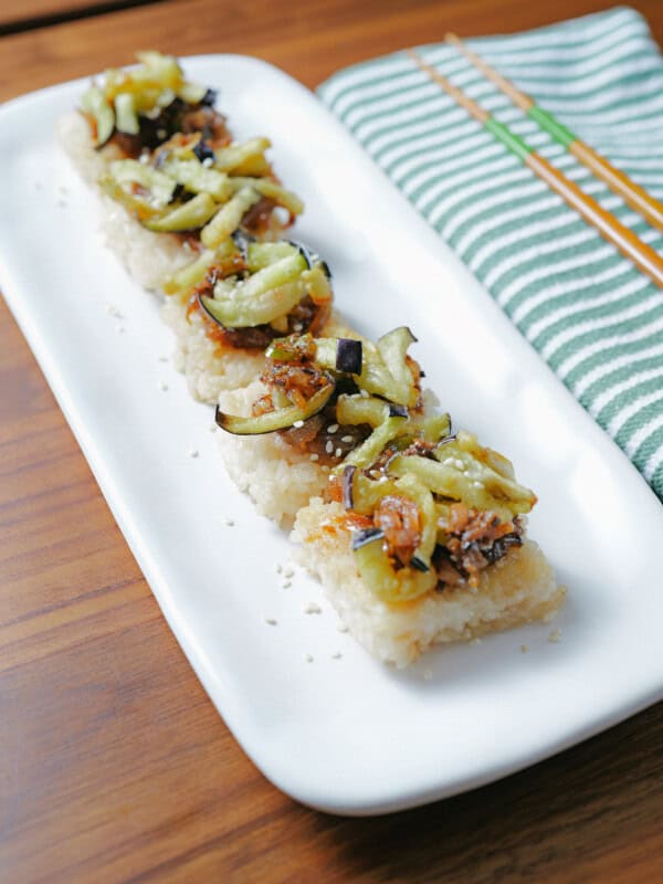 A rectangular white plate with four pieces of crispy rice topped with a vegetable mixture, placed on a wooden table next to a green and white striped napkin and a pair of chopsticks.