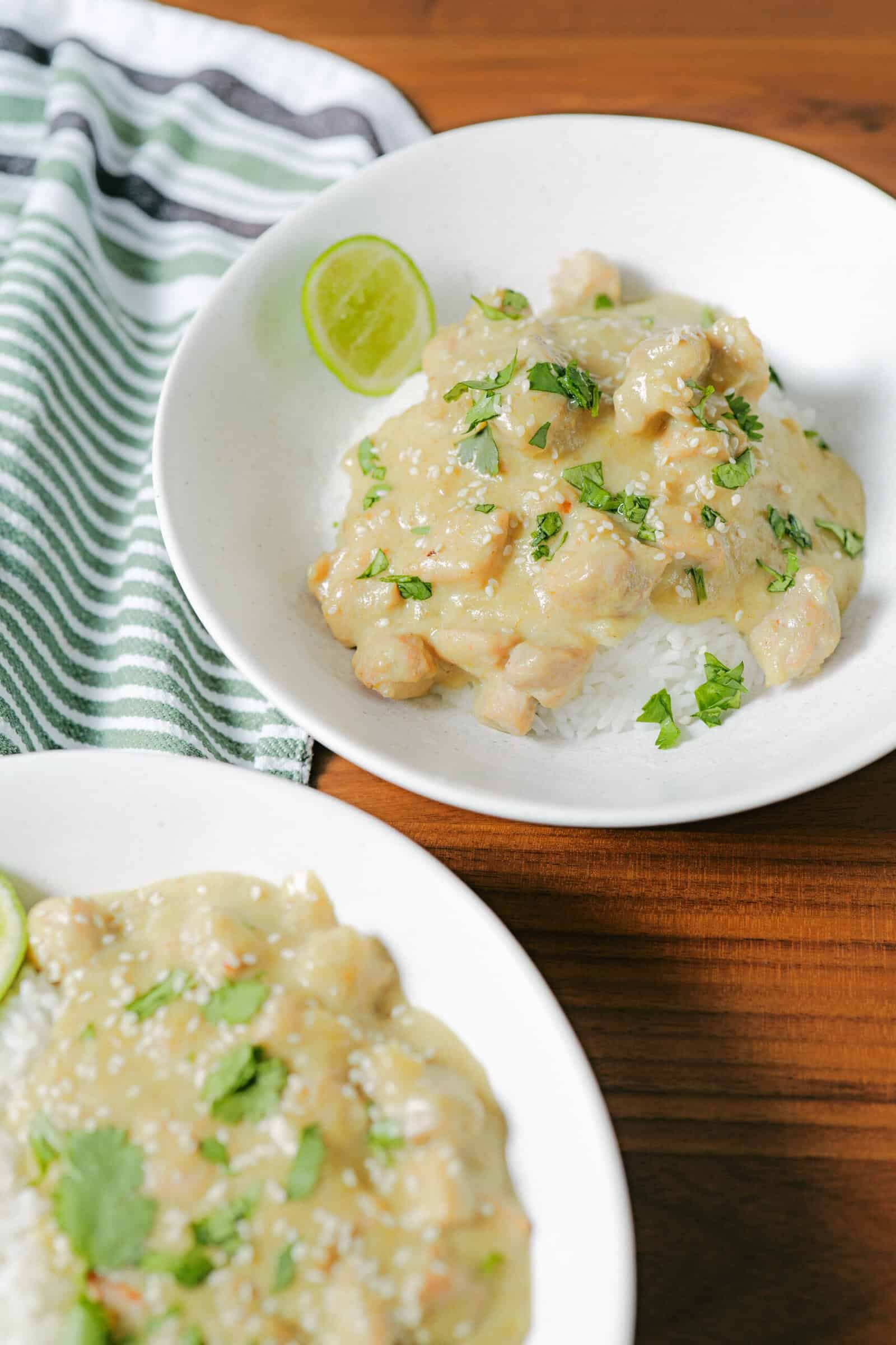 Two white bowls filled with white rice topped with creamy chicken curry, garnished with chopped cilantro and lime slices, are set on a wooden table beside a green and white striped cloth.