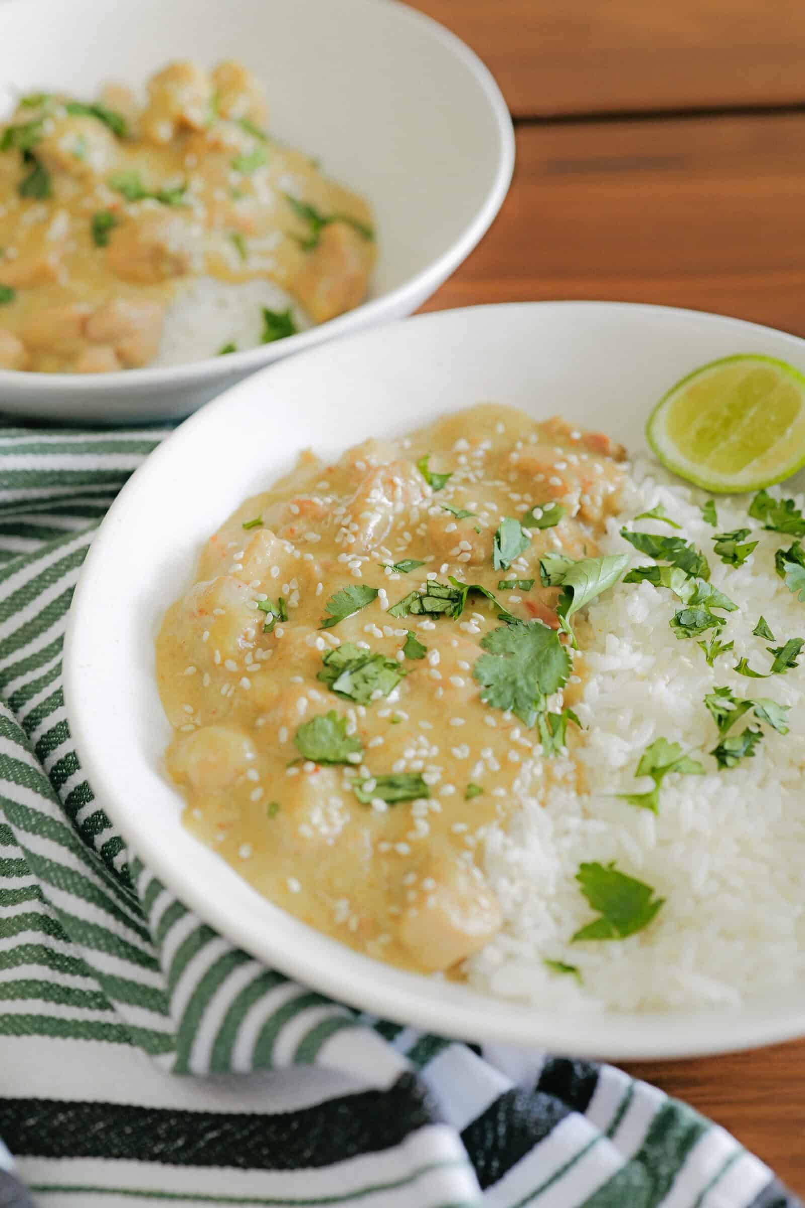 A white bowl filled with white rice and creamy curry topped with chopped cilantro, sesame seeds, and a lime wedge. Another similar bowl is blurred in the background, with a striped green and white cloth nearby.