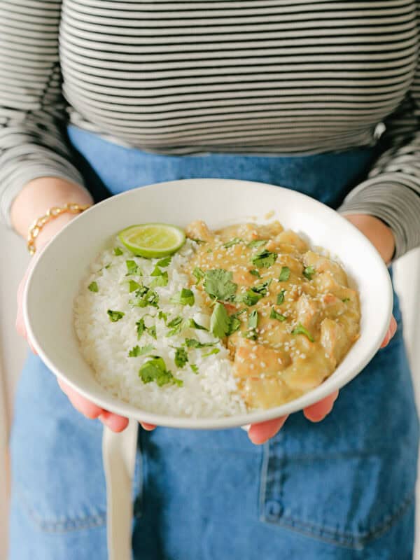 A person in a striped shirt and blue apron holds a bowl of white rice topped with saucy chicken, cilantro, and a lime wedge.