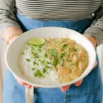 A person in a striped shirt and blue apron holds a bowl with white rice, creamy curry, fresh cilantro, and a lime wedge.