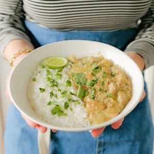 A person in a striped shirt and blue apron holds a bowl with white rice, creamy curry, fresh cilantro, and a lime wedge.