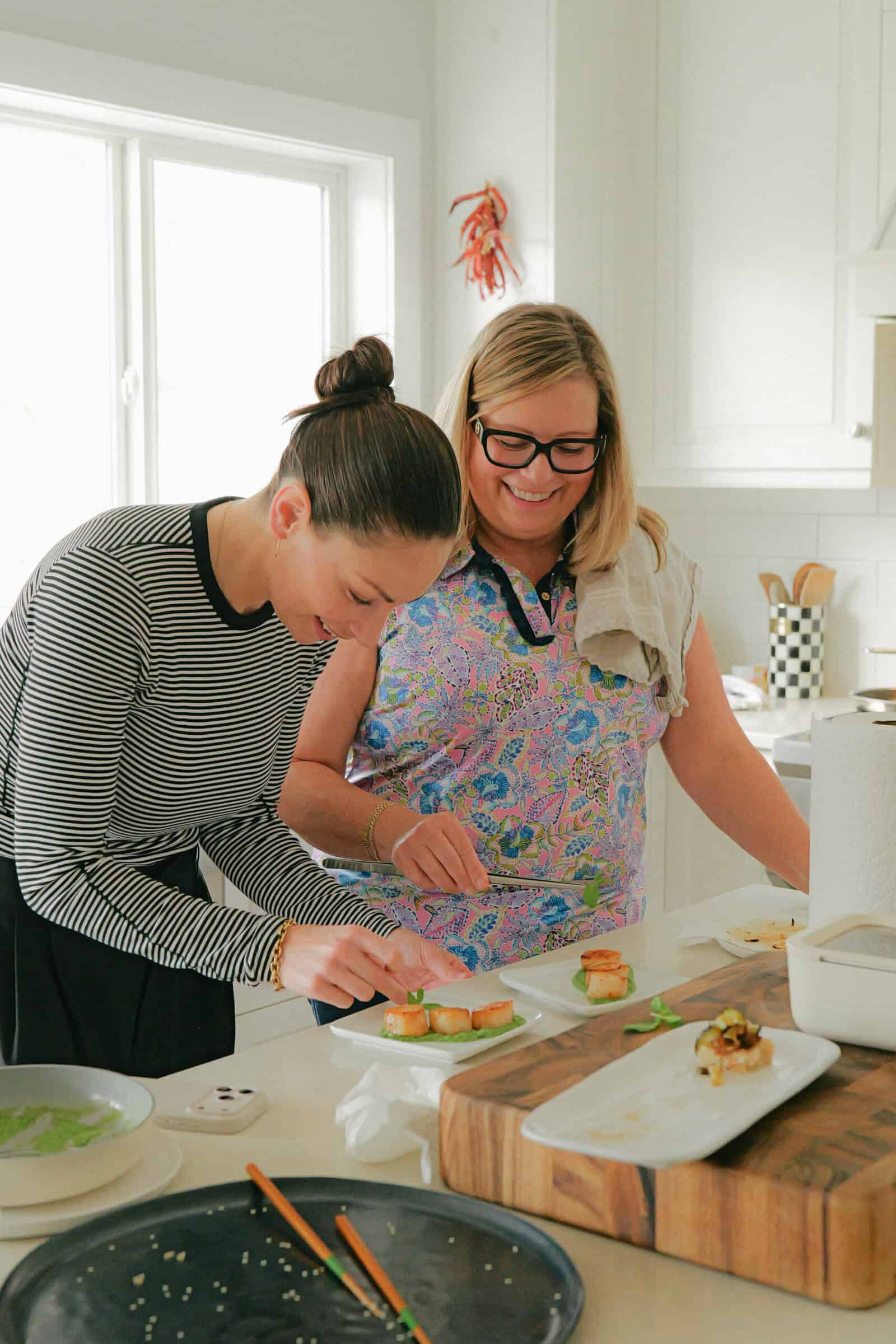 Two women stand in a bright kitchen preparing food together, smiling and plating small dishes on a counter. The scene is casual and friendly, with sunlight streaming through a window in the background.