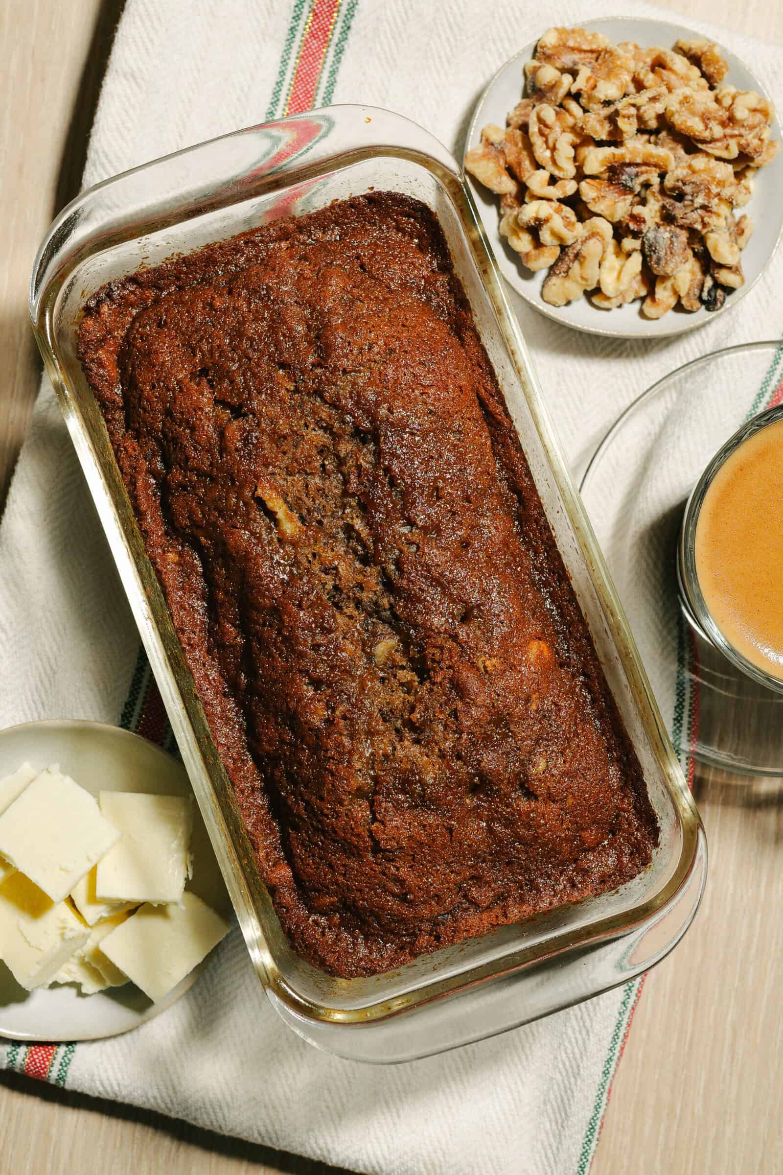 A loaf of banana bread in a glass baking dish on a striped cloth, with a plate of butter slices, walnuts, and coffee nearby—perfect inspiration for trying your own Date Nut Bread Recipe.