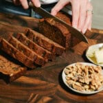 A person slices a loaf of Date Nut Bread on a wooden cutting board. Several slices are cut, with a small plate of walnuts and butter nearby. The person wears a striped shirt and a bracelet.