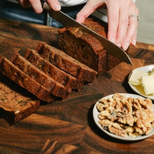 A person slices a loaf of Date Nut Bread on a wooden cutting board. Several slices are cut, with a small plate of walnuts and butter nearby. The person wears a striped shirt and a bracelet.