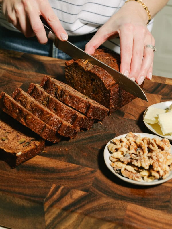 A person slices a loaf of Date Nut Bread on a wooden cutting board. Several slices are cut, with a small plate of walnuts and butter nearby. The person wears a striped shirt and a bracelet.