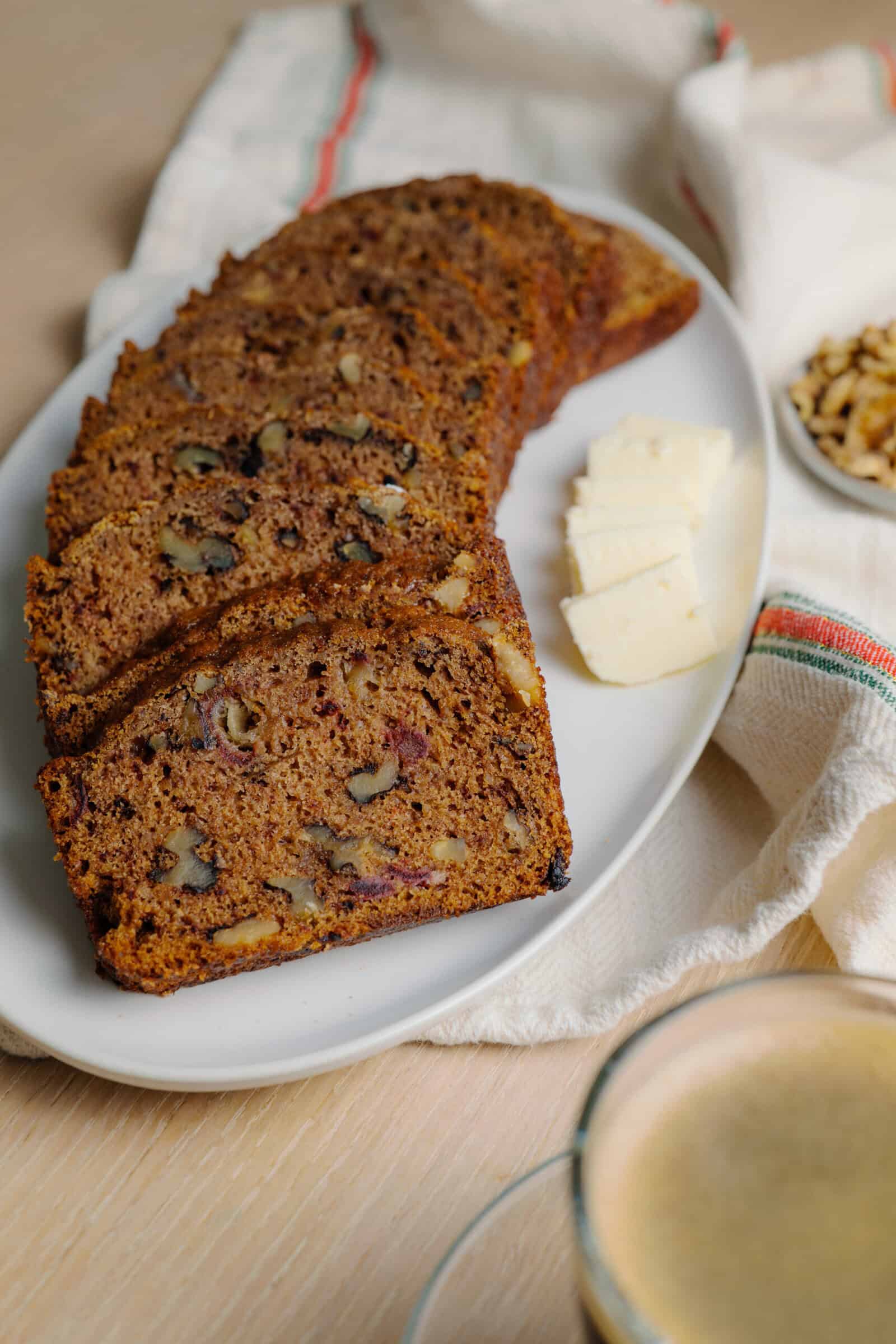 A white plate with several slices of banana bread containing walnuts, three slices of butter, a bowl of chopped walnuts, and a cup of coffee on a light wooden surface evokes the cozy feel of a classic Date Nut Bread recipe experience.