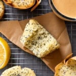 A close-up of a Lemon Poppy Seed Muffin with a bite taken out, placed on brown paper atop a cooling rack. More Lemon Poppy Seed Muffins, a half lemon, and a cup of coffee complete this inviting scene—perfect for your next Lemon Poppy Seed Muffins Recipe.