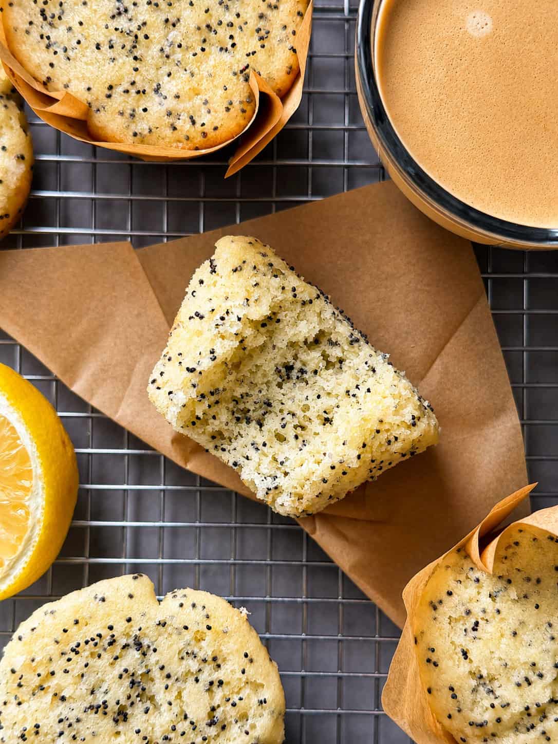 A close-up of a Lemon Poppy Seed Muffin with a bite taken out, placed on brown paper atop a cooling rack. More Lemon Poppy Seed Muffins, a half lemon, and a cup of coffee complete this inviting scene—perfect for your next Lemon Poppy Seed Muffins Recipe.