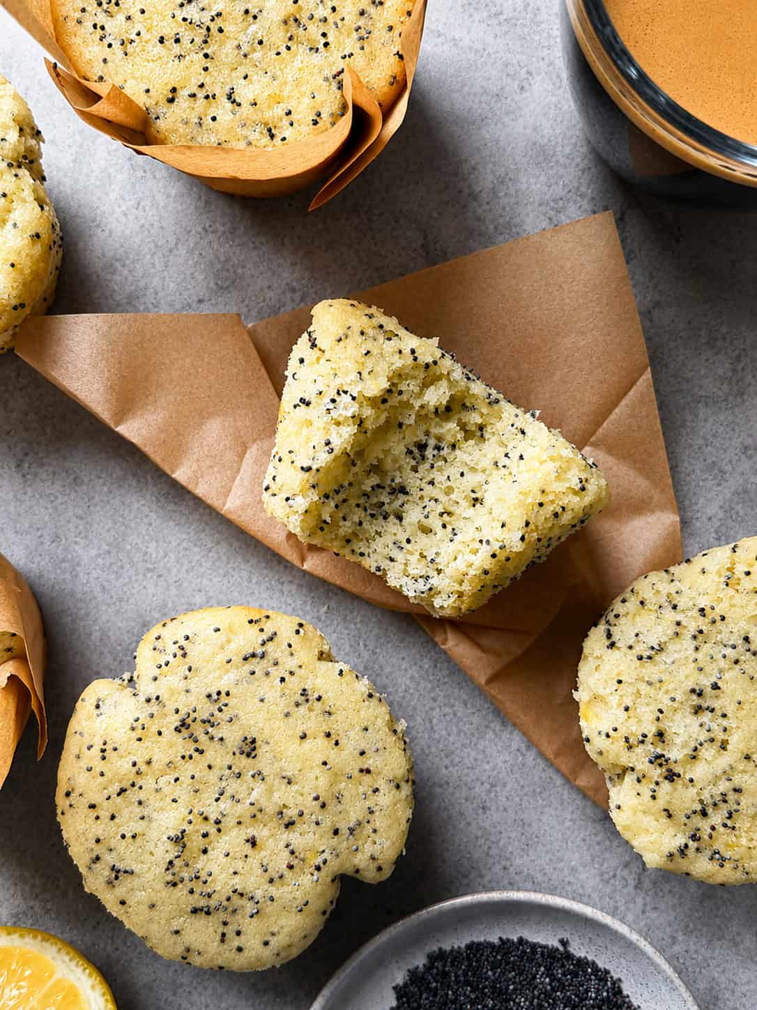 Four Lemon Poppy Seed Muffins, one split in half to reveal the moist texture, sit on brown parchment paper atop a gray surface. Nearby are a bowl of poppy seeds and a glass of coffee—perfect for trying your favorite Lemon Poppy Seed Muffins recipe.
