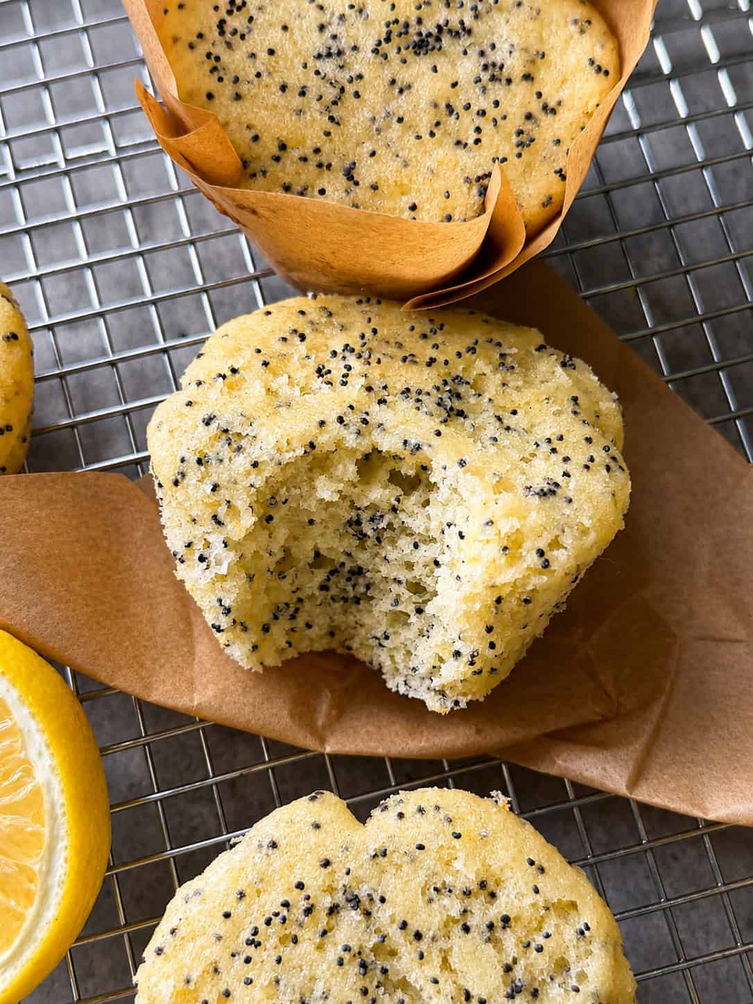 A close-up of Lemon Poppy Seed Muffins on a cooling rack, one partially unwrapped with a bite taken out, showing the soft, speckled interior. A lemon wedge hints at the zesty flavor of this Lemon Poppy Seed Muffins Recipe.