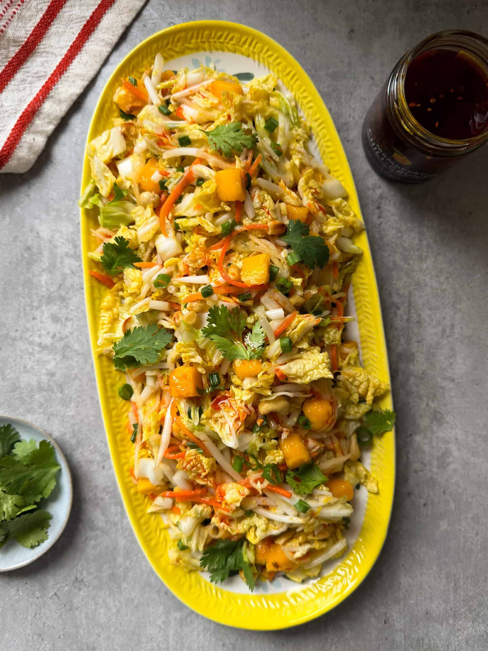 A yellow oval platter holds a vibrant Crunchy Asian Slaw with napa cabbage, carrots, mango chunks, cilantro, and a light dressing. Nearby are a jar of sauce, cilantro on a small plate, and a striped cloth on the gray surface.