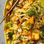 A vibrant Crunchy Asian Slaw with shredded cabbage, carrots, mango chunks, cilantro, green onions, and peanuts on a yellow platter, garnished with fresh herbs. Chopsticks rest on the side of the dish.