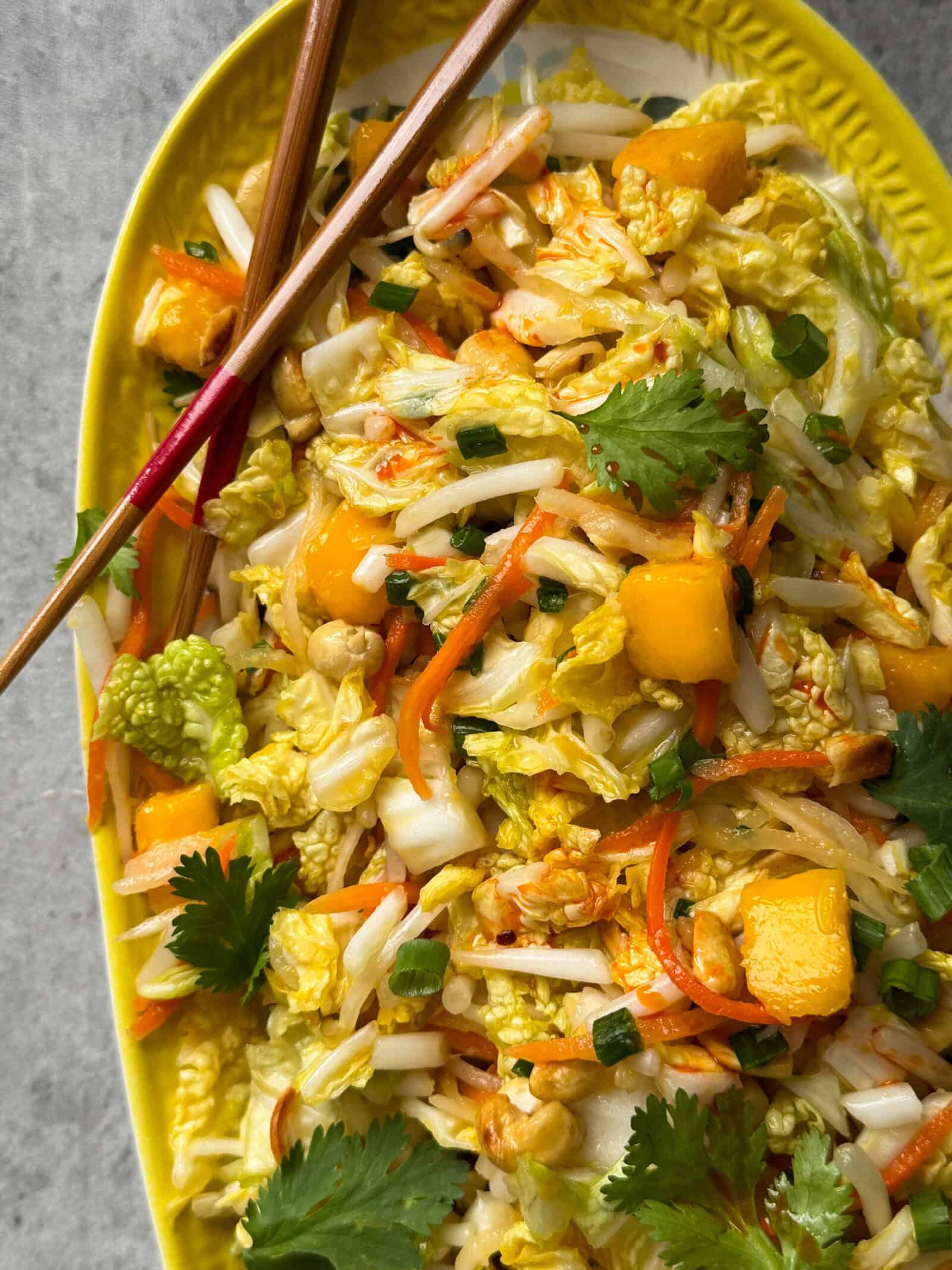 A vibrant Crunchy Asian Slaw with shredded cabbage, carrots, mango chunks, cilantro, green onions, and peanuts on a yellow platter, garnished with fresh herbs. Chopsticks rest on the side of the dish.