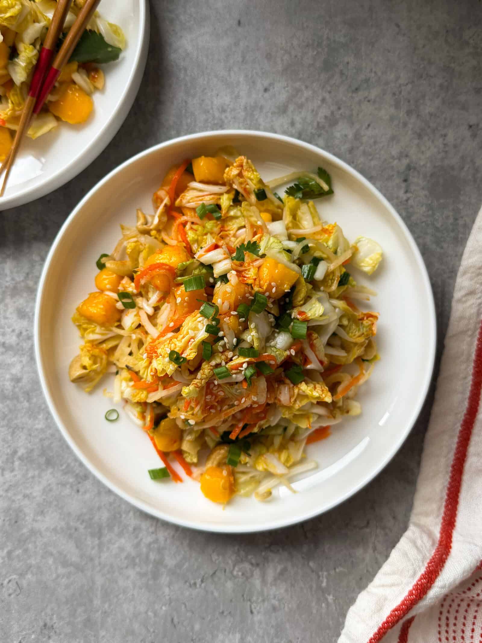 A white plate with a Crunchy Asian Slaw featuring napa cabbage, shredded carrots, green onions, mandarin orange segments, and sesame seeds sits on a gray surface with a striped towel and chopsticks nearby.