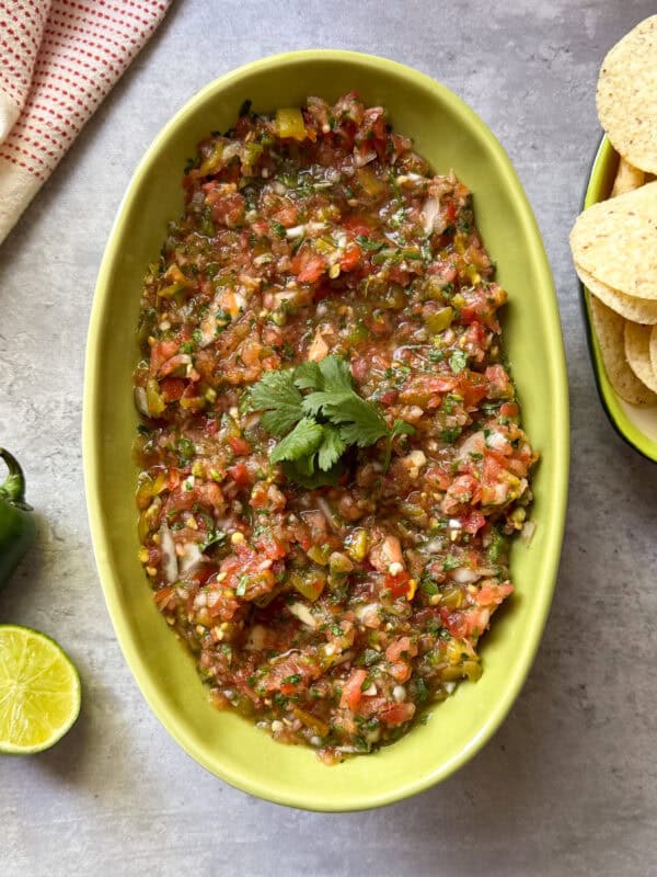 A green oval dish filled with chunky fresh salsa recipe, garnished with cilantro, sits on a gray surface next to tortilla chips, a halved lime, a green pepper, and a red-and-white cloth.