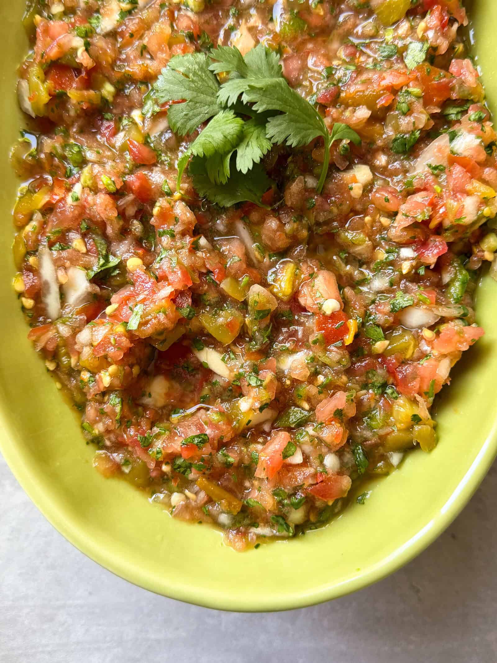 Fresh tomato salsa with chopped tomatoes, onions, cilantro, and green peppers in a green bowl—this homemade salsa recipe is garnished with a sprig of cilantro on top.