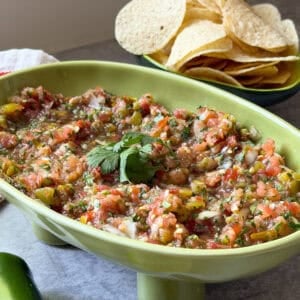 A green dish filled with a fresh salsa recipe garnished with cilantro, with a plate of tortilla chips in the background. Sliced lime and a jalapeño are on the table beside this vibrant homemade salsa recipe.