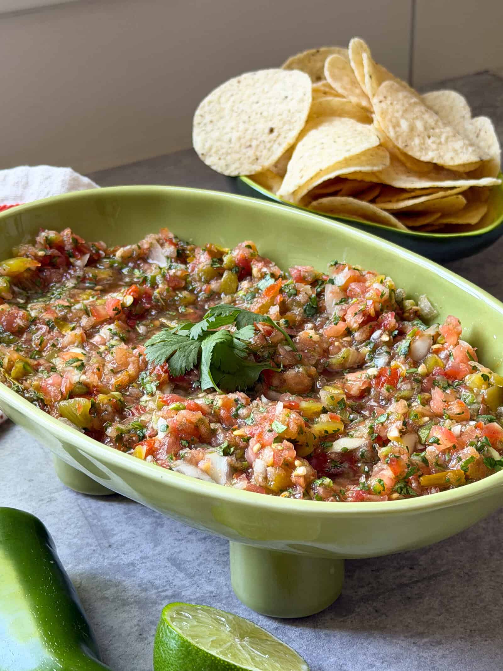 A green dish filled with a fresh salsa recipe garnished with cilantro, with a plate of tortilla chips in the background. Sliced lime and a jalapeño are on the table beside this vibrant homemade salsa recipe.