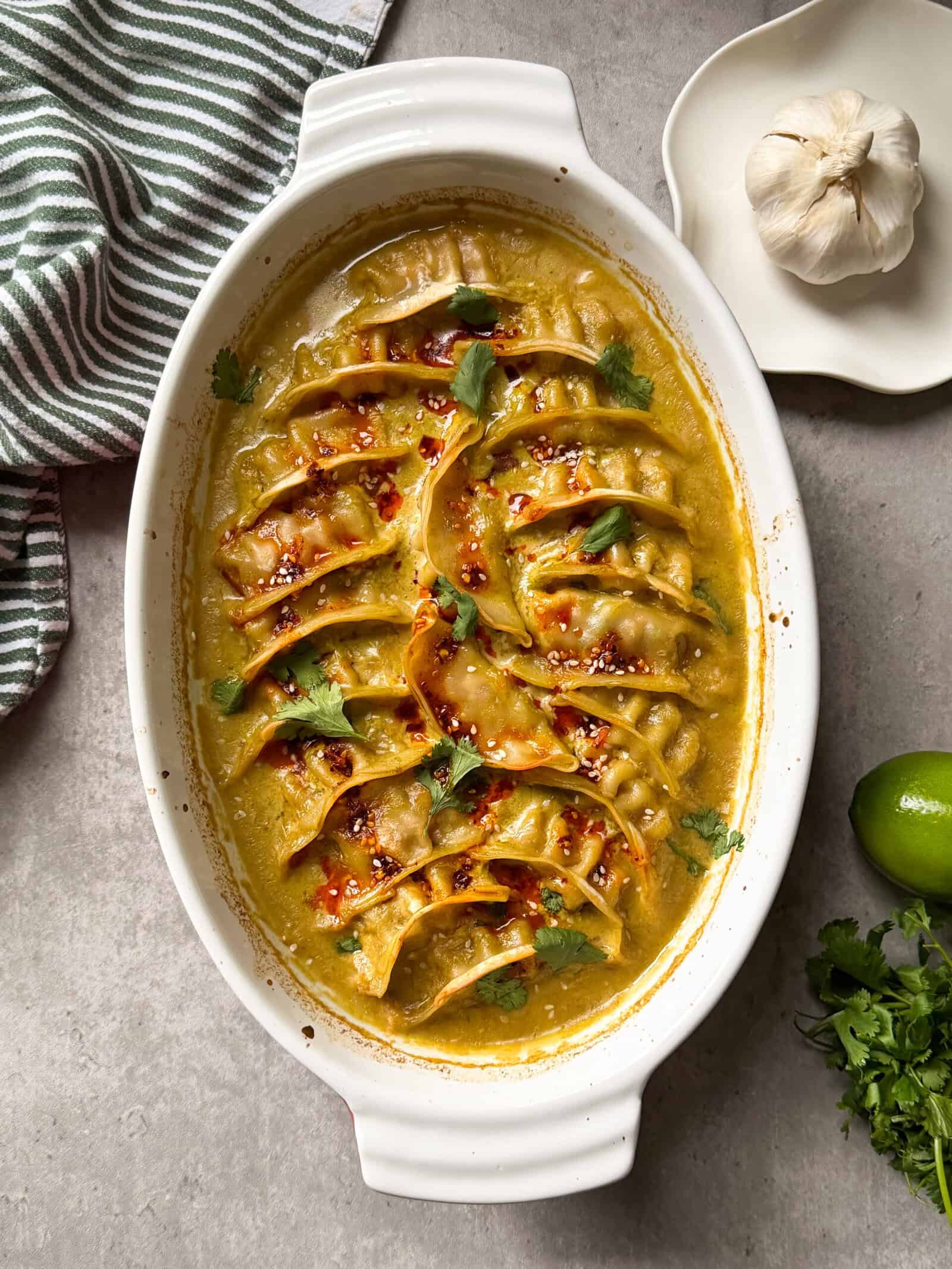 A white oval dish filled with dumplings in a yellow curry sauce, garnished with cilantro—this Dumpling Bake Recipe is surrounded by a garlic bulb, lime, fresh cilantro, and a striped cloth napkin on a gray surface.