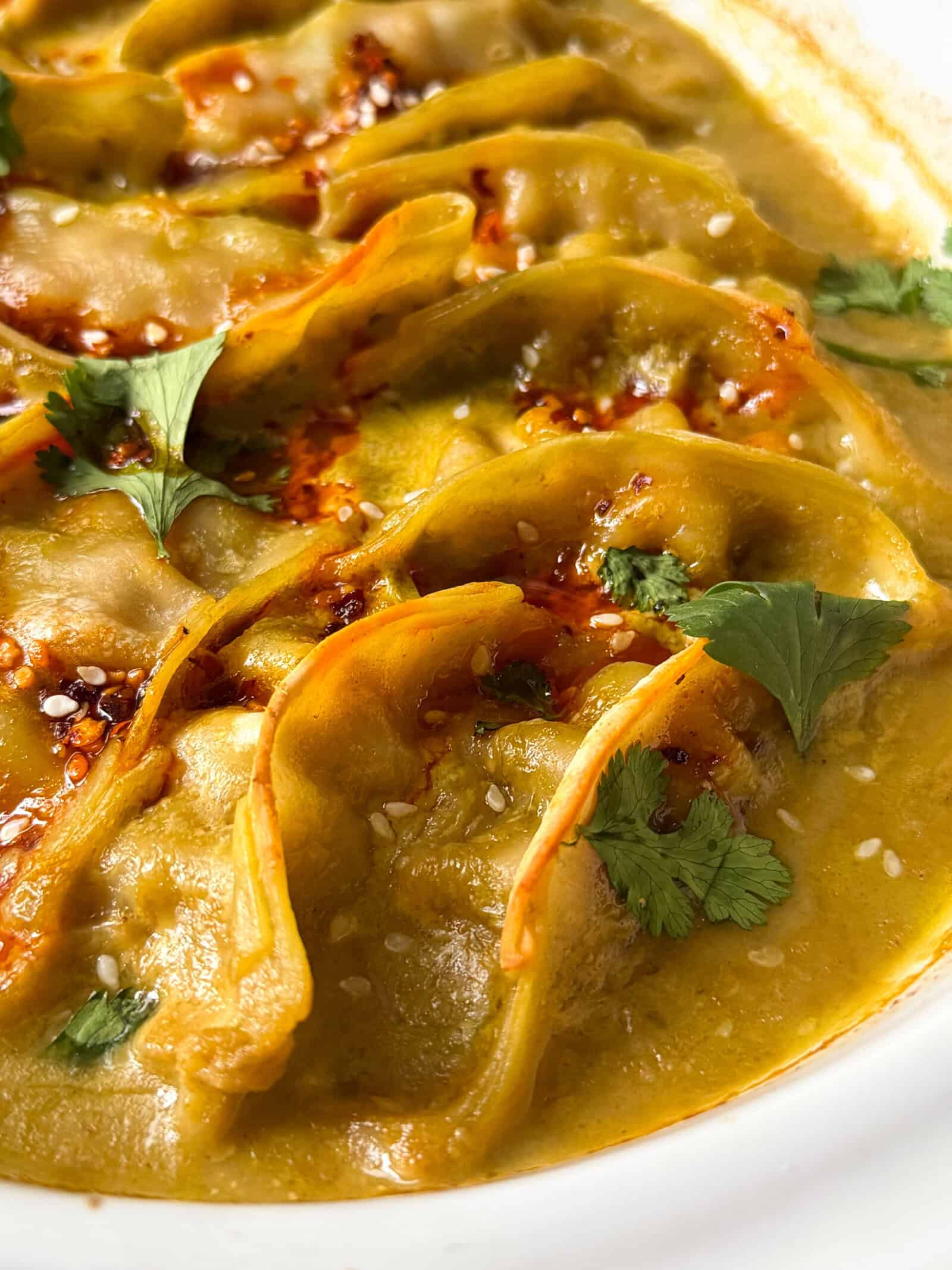 Close-up of Viral Dumpling Bake: steamed dumplings in a golden, spicy sauce, garnished with cilantro leaves, sesame seeds, and a drizzle of chili oil.
