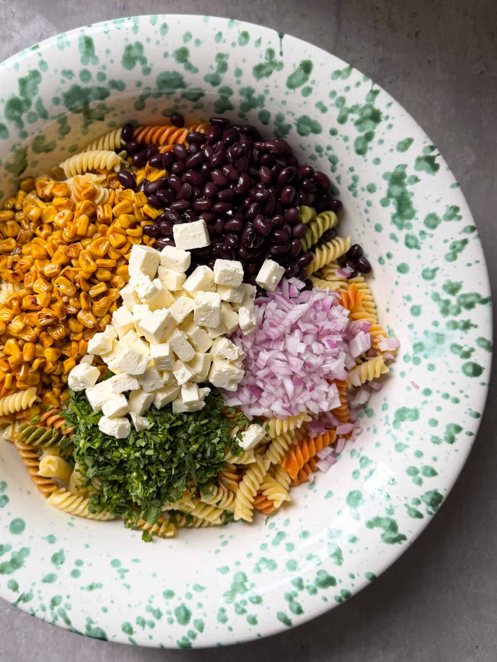 A vibrant Mexican Street Corn Pasta Salad Recipe featuring colorful rotini pasta, black beans, roasted corn, diced tofu, red onion, and fresh herbs arranged on a white plate with green speckles.