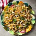 A colorful plate of Mexican Street Corn Pasta Salad with black beans, corn, feta cheese, chopped vegetables, and fresh cilantro, served on a decorative leaf-shaped dish beside a red and white striped cloth.