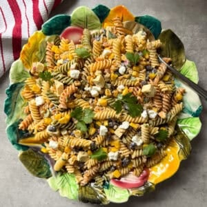 A colorful plate of Mexican Street Corn Pasta Salad with black beans, corn, feta cheese, chopped vegetables, and fresh cilantro, served on a decorative leaf-shaped dish beside a red and white striped cloth.