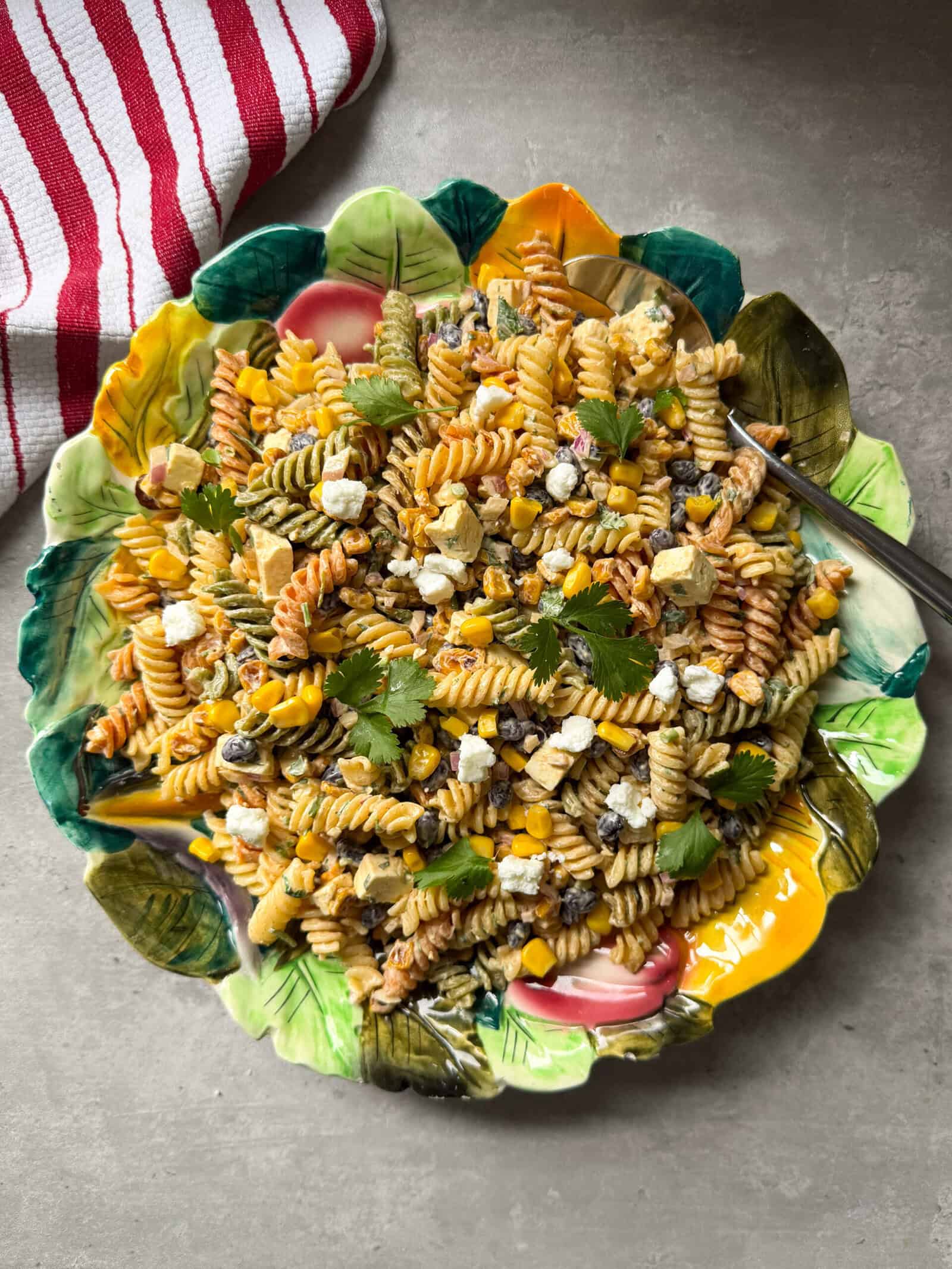 A colorful plate of Mexican Street Corn Pasta Salad with black beans, corn, feta cheese, chopped vegetables, and fresh cilantro, served on a decorative leaf-shaped dish beside a red and white striped cloth.