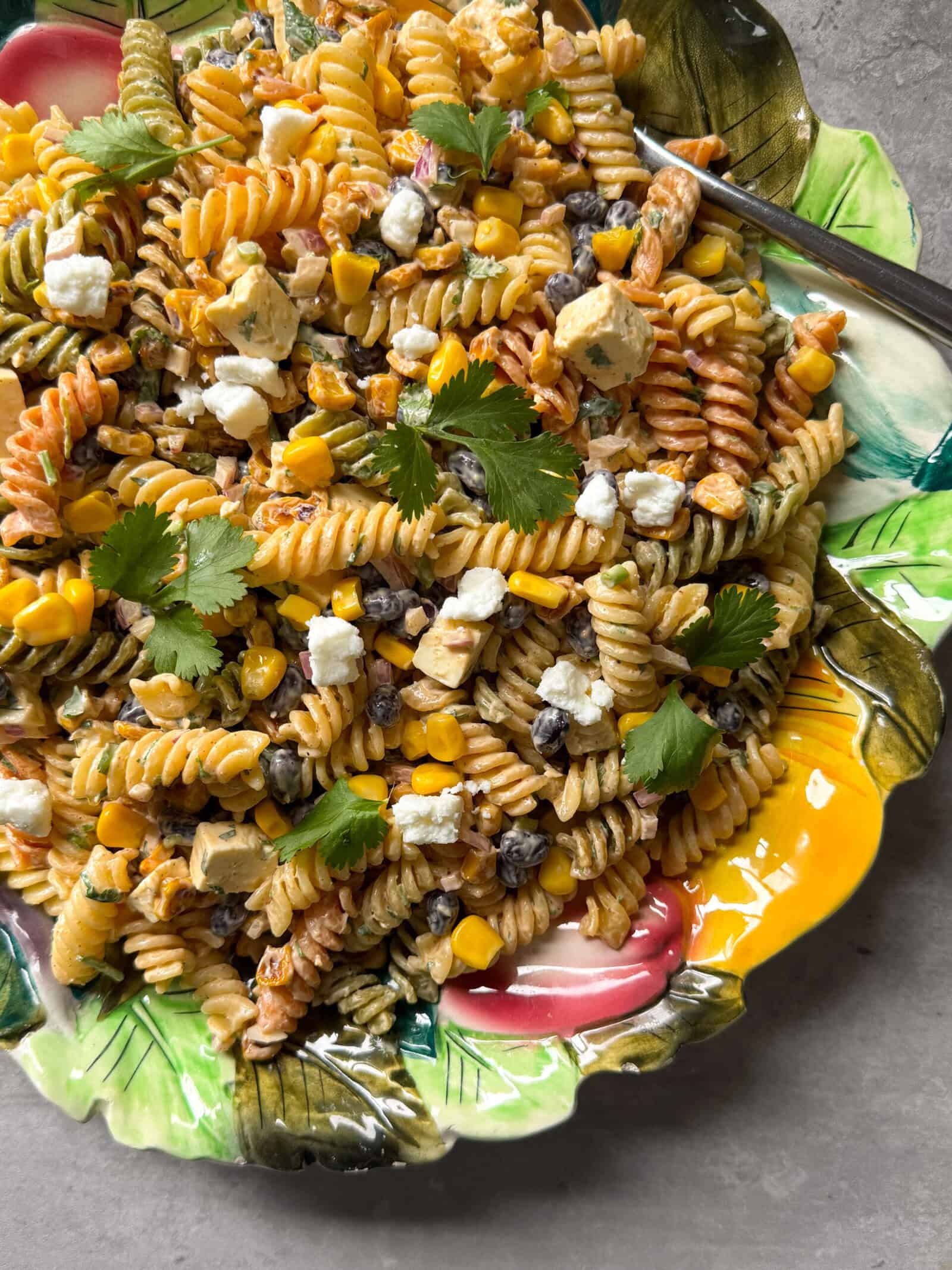 A vibrant plate of Mexican Street Corn Pasta Salad with rotini, corn, black beans, cilantro, and crumbled cheese sits on a floral dish, with a fork on the side—perfect inspiration for your next Pasta Salad Recipes collection.