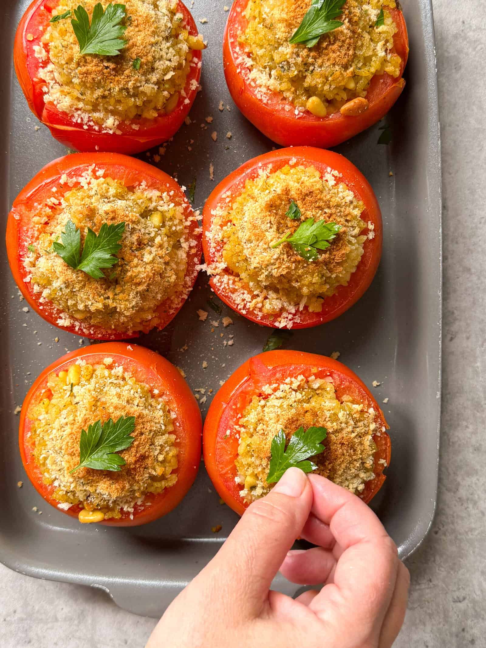 A hand places parsley on top of baked stuffed tomatoes filled with a flavorful breadcrumb and corn mixture, arranged neatly in a baking dish—an irresistible stuffed tomatoes recipe.