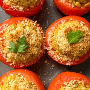 Six stuffed tomatoes, made with our classic stuffed tomatoes recipe and filled with a golden crumb mixture, are garnished with fresh parsley and arranged close together on a dark surface.