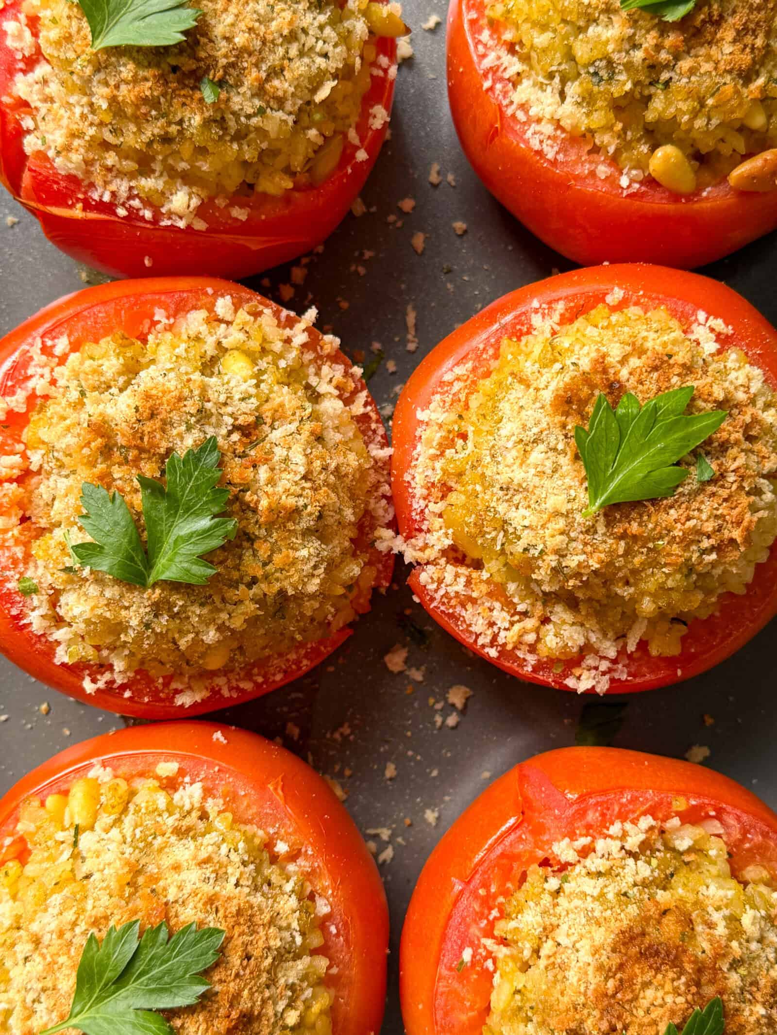 Six stuffed tomatoes, made with our classic stuffed tomatoes recipe and filled with a golden crumb mixture, are garnished with fresh parsley and arranged close together on a dark surface.
