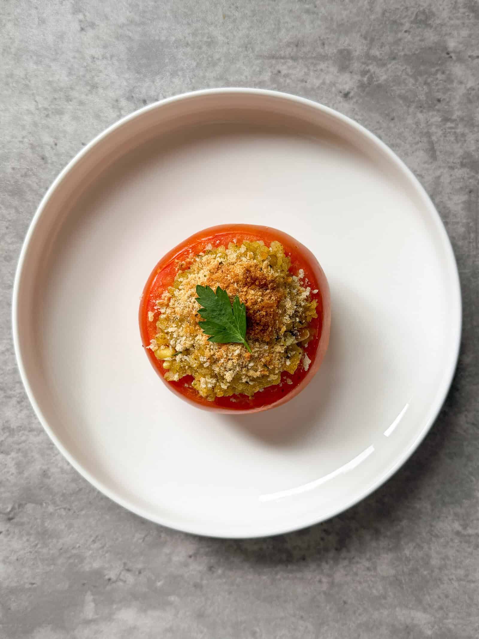 A delicious stuffed tomato, inspired by a classic stuffed tomatoes recipe, is topped with golden breadcrumbs and fresh herbs. It sits on a white plate, viewed from above on a gray countertop.