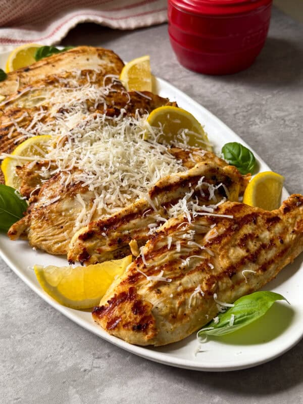 Grilled Lemon Caesar Yogurt Marinated Chicken breasts topped with grated cheese are arranged on a white platter, garnished with fresh basil leaves and lemon wedges. A red container is in the background on a gray surface.