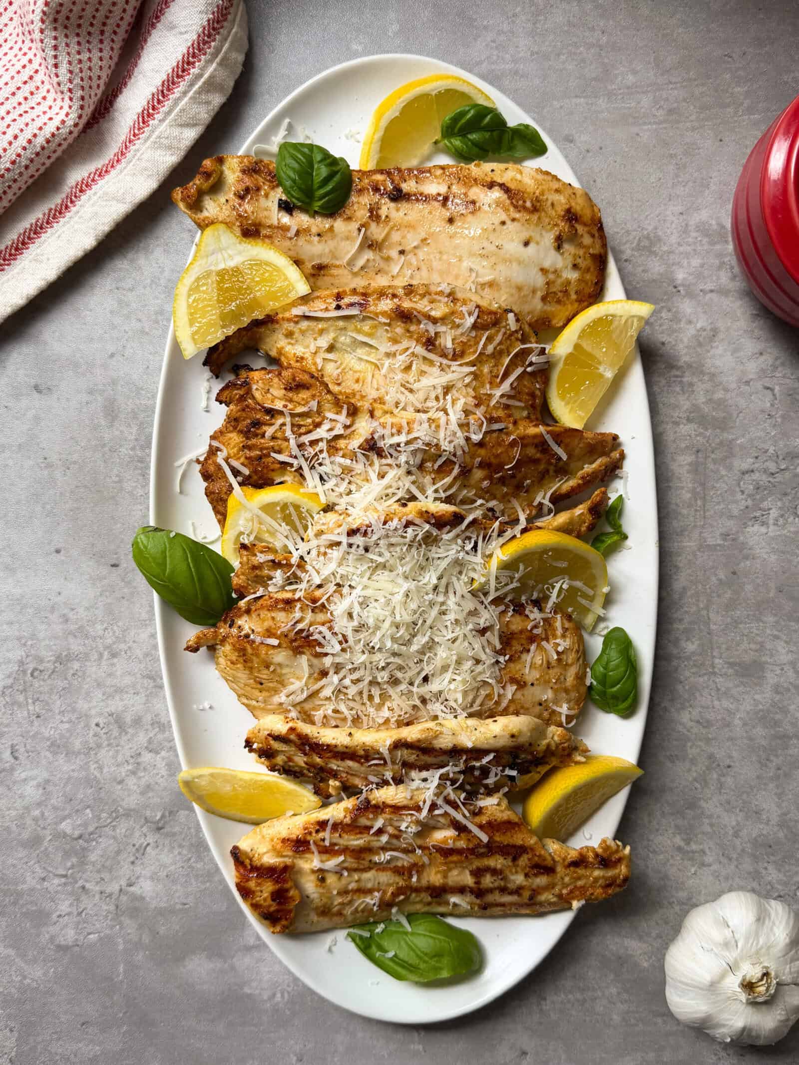 Grilled Lemon Caesar Yogurt Marinated Chicken breasts arranged on an oval white platter, topped with grated cheese and garnished with lemon wedges and fresh basil, sit on a gray stone surface. A garlic bulb and red cloth are nearby.