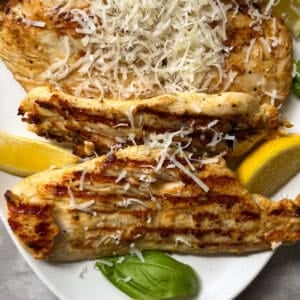 Grilled Lemon Caesar Yogurt Marinated Chicken breasts topped with shredded Parmesan, served on a white plate with lemon wedges and fresh basil leaves.