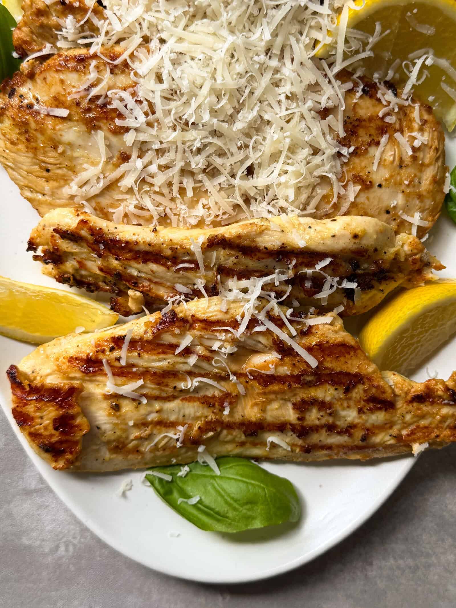 Grilled Lemon Caesar Yogurt Marinated Chicken breasts topped with shredded Parmesan, served on a white plate with lemon wedges and fresh basil leaves.