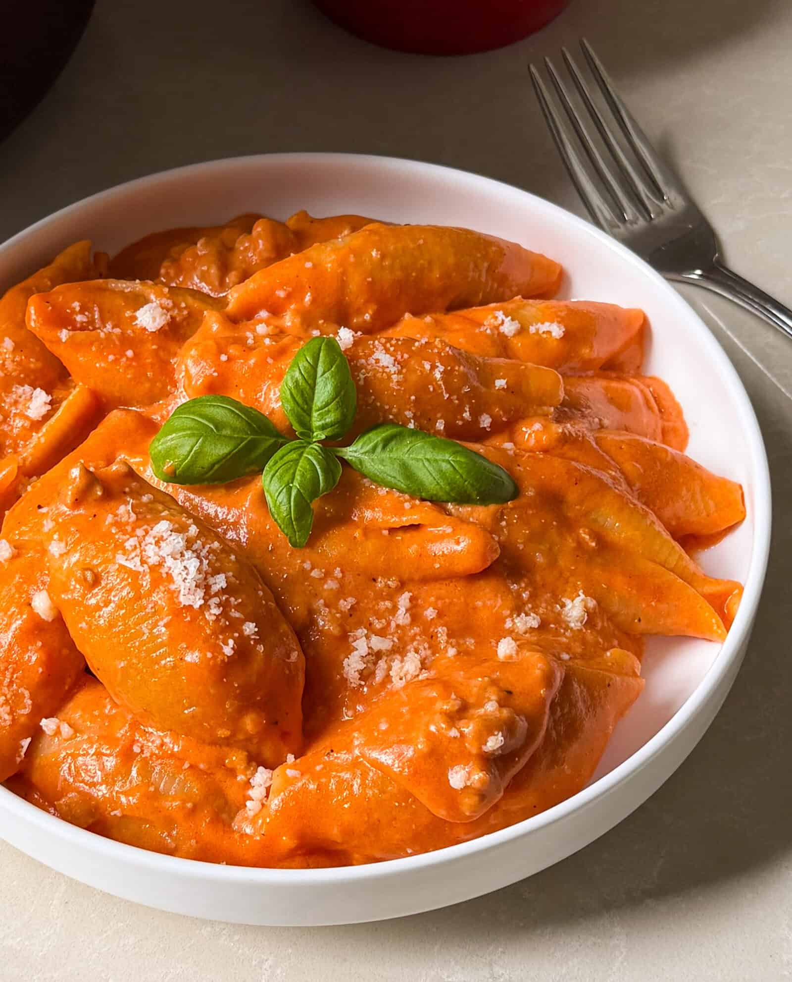 A white bowl filled with pasta shells in creamy tomato sauce, topped with grated cheese and a basil garnish, sits on a light surface next to a fork—an inviting unstuffed shells recipe that’s both simple and delicious.