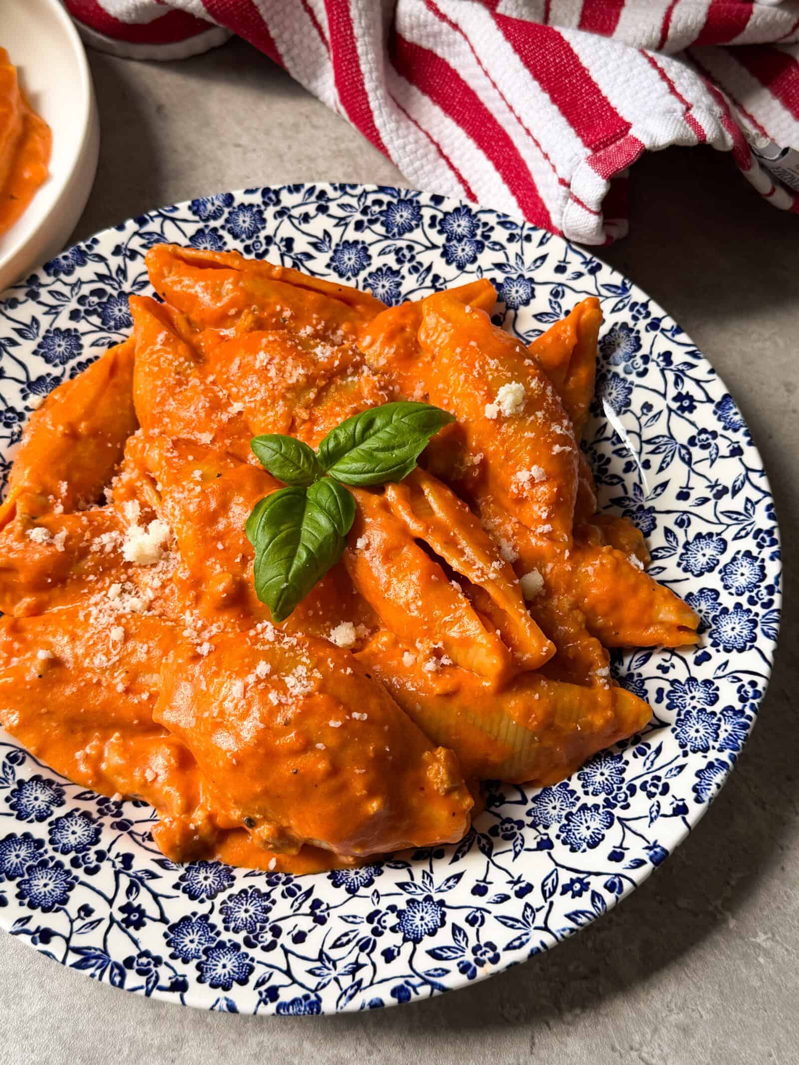 Penne pasta coated in creamy tomato sauce, reminiscent of a saucy unstuffed shells recipe, is garnished with grated cheese and a fresh basil sprig, served on a blue and white floral plate. A red and white striped towel is in the background.