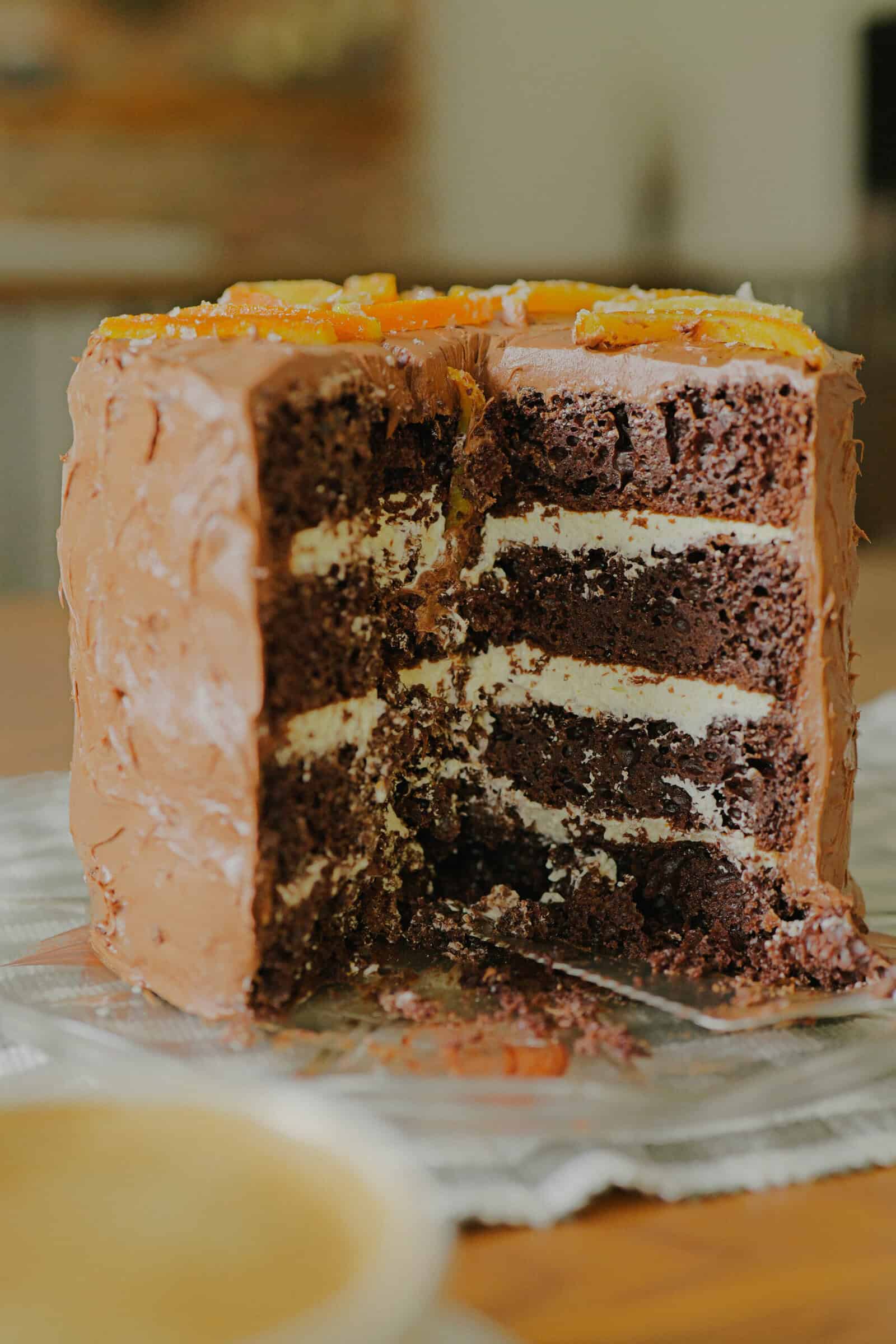 A partially sliced, tall chocolate layer cake with chocolate frosting and cream filling sits on a table. The top is decorated with pieces of fruit. A blurred cup is visible in the foreground.