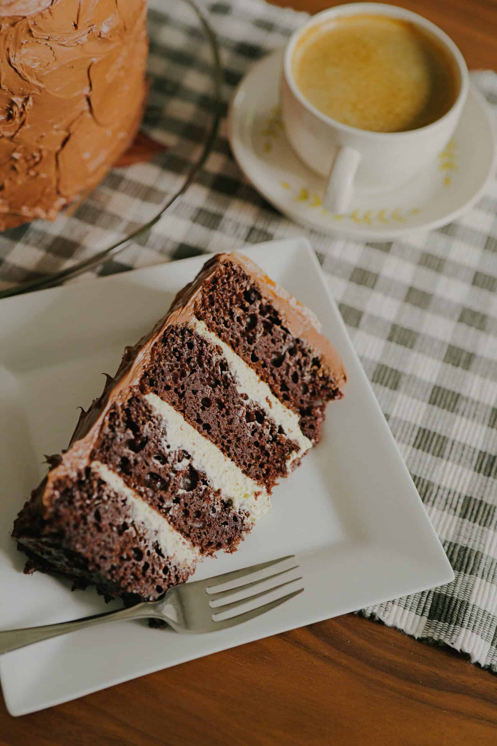 A slice of layered chocolate and cream cake on a white plate with a fork, next to a cup of coffee on a plaid cloth and a wooden table.