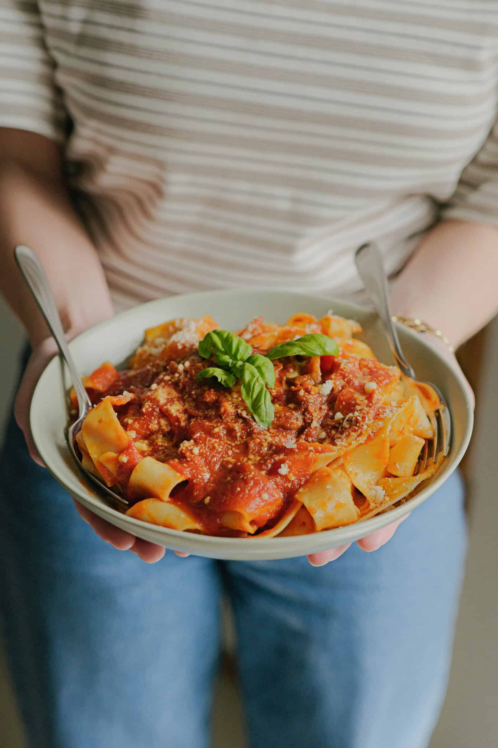A person in a striped shirt and jeans holds a bowl of pasta topped with tomato sauce, ground meat, grated cheese, and a basil garnish, with a fork and spoon in the bowl.