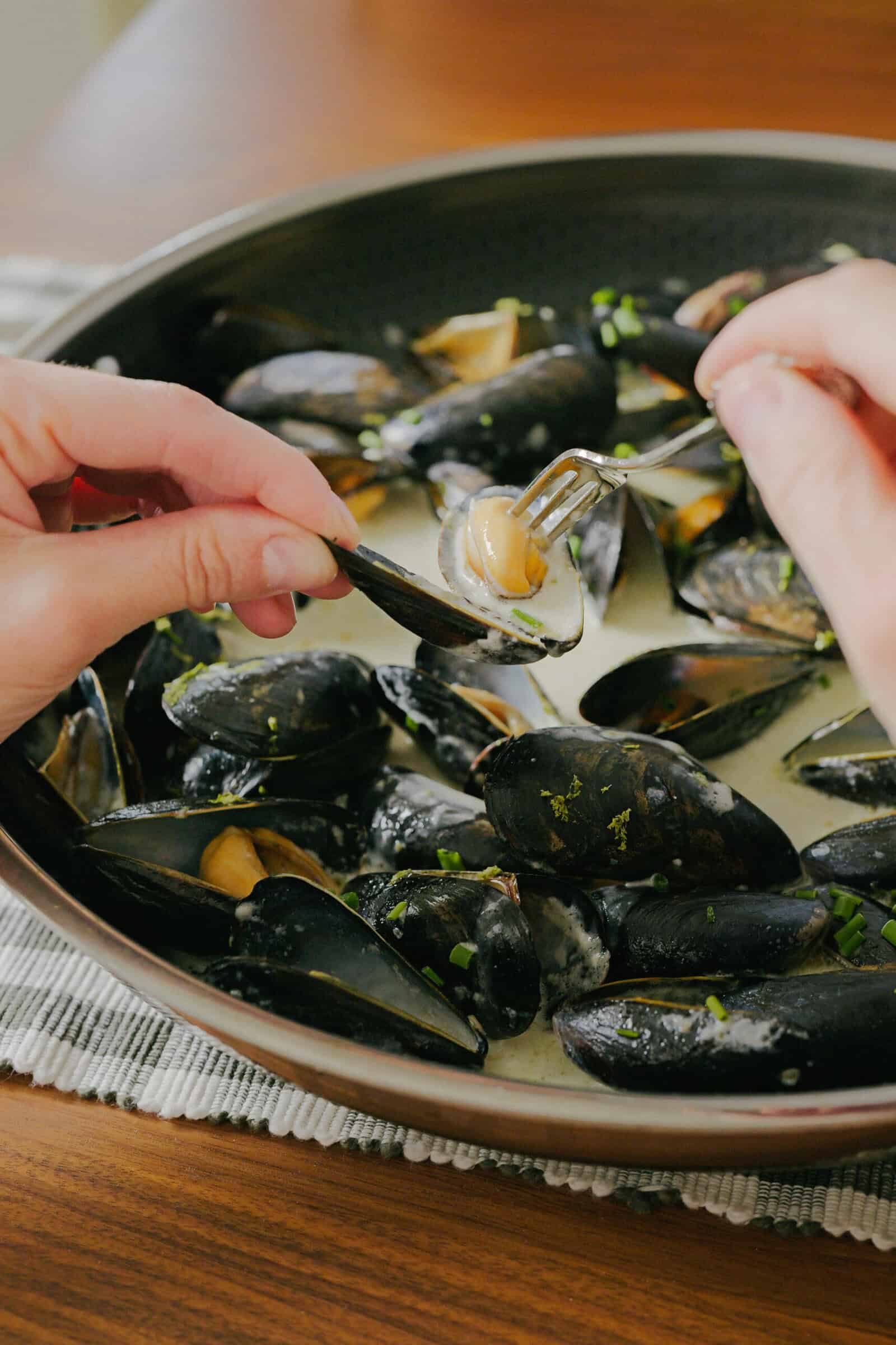 A person uses a fork to remove a cooked mussel from its shell over a pan filled with steamed mussels in a creamy sauce, garnished with chopped herbs.