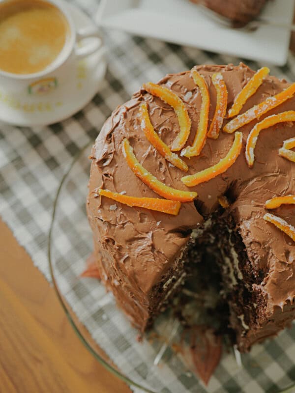 A chocolate layer cake with orange slices on top sits on a glass plate, with a slice missing. Next to it is a cup of coffee on a checkered cloth.