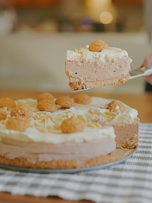 A hand lifts a slice of creamy, layered cake topped with whipped cream and small round cookies from a whole cake on a table with a checkered cloth.