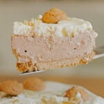 A slice of creamy, light brown cheesecake topped with whipped cream, a small cookie, and almond slivers, held above the rest of the cake.