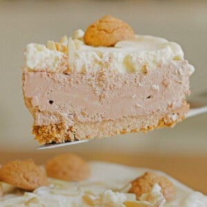 A slice of creamy, light brown cheesecake topped with whipped cream, a small cookie, and almond slivers, held above the rest of the cake.
